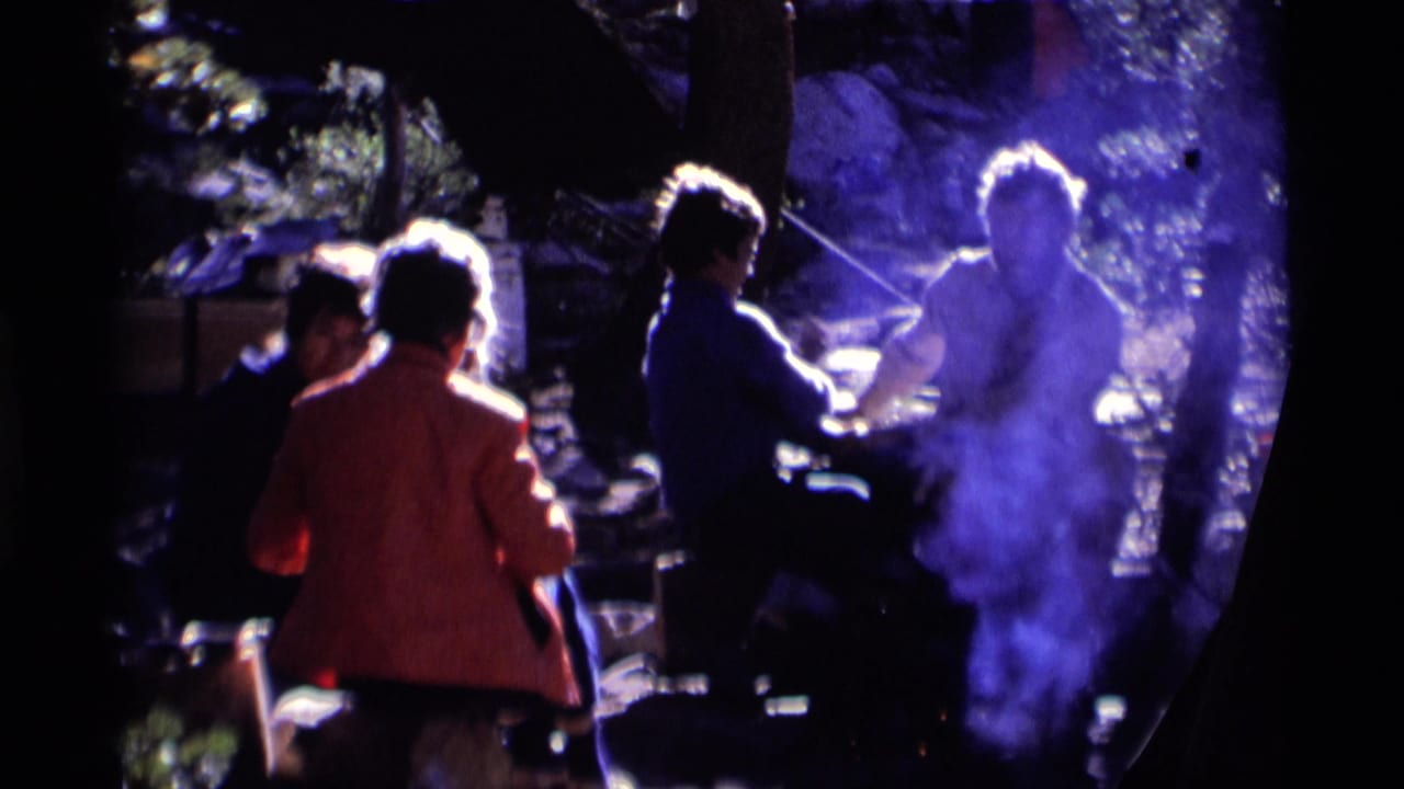 Backlit 1975 home-movie frame of adults gathered near a smoking campfire in the Minaret Wilderness woods.