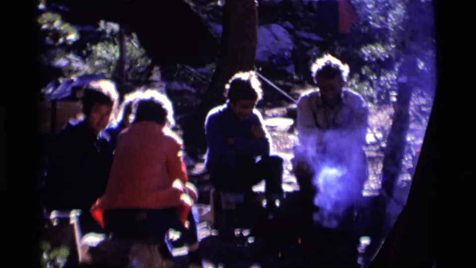 A 1975 8mm camping scene shows adults in casual jackets seated beside a smoky fire in a wooded California campsite.