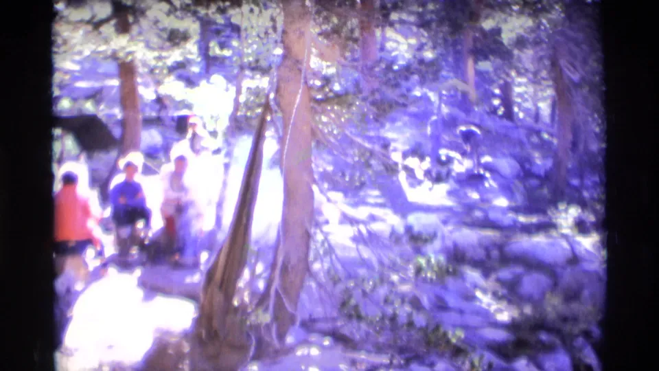 Wide 8mm frame from the Minaret Wilderness campsite, with figures near a bright campfire amid trees, rocks, and smoke.