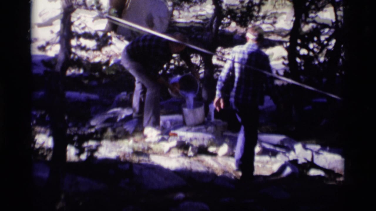In 1975 Minaret Wilderness footage, two men in plaid shirts stand among pine trees and rocks near buckets and a small stone basin.