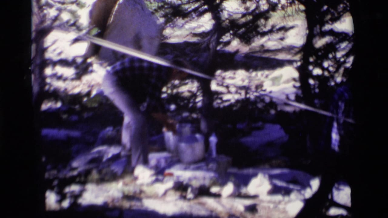 A man bends near a bucket and stone basin in the California wilderness, with rocks, tree shadows, and a line crossing the frame.