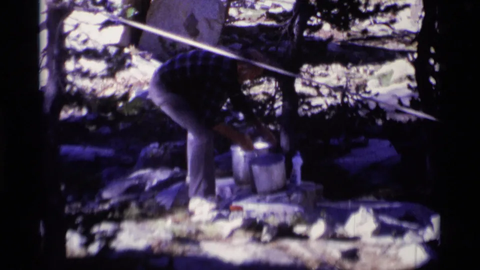 A man in a plaid shirt appears to pour or handle water at a stone basin in the Minaret Wilderness, surrounded by rocks and trees.