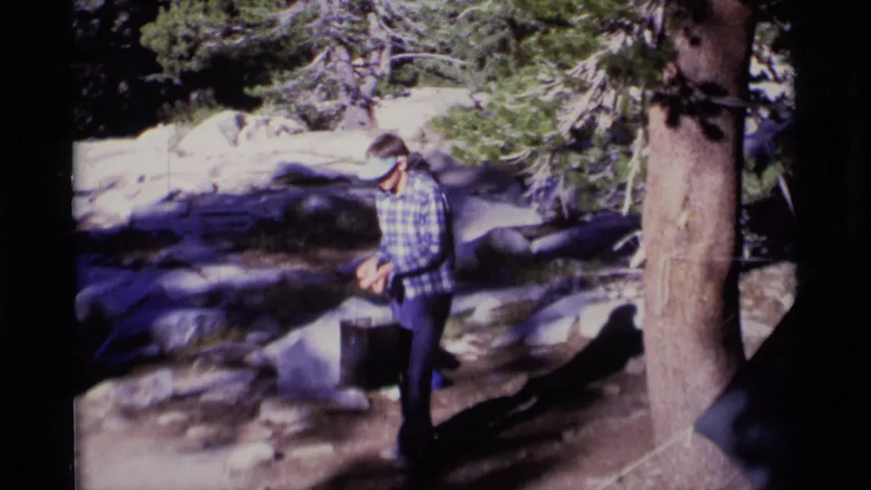 A man in a plaid shirt carries a dark bucket through a rocky pine forest in 1975 Minaret Wilderness home-movie footage.