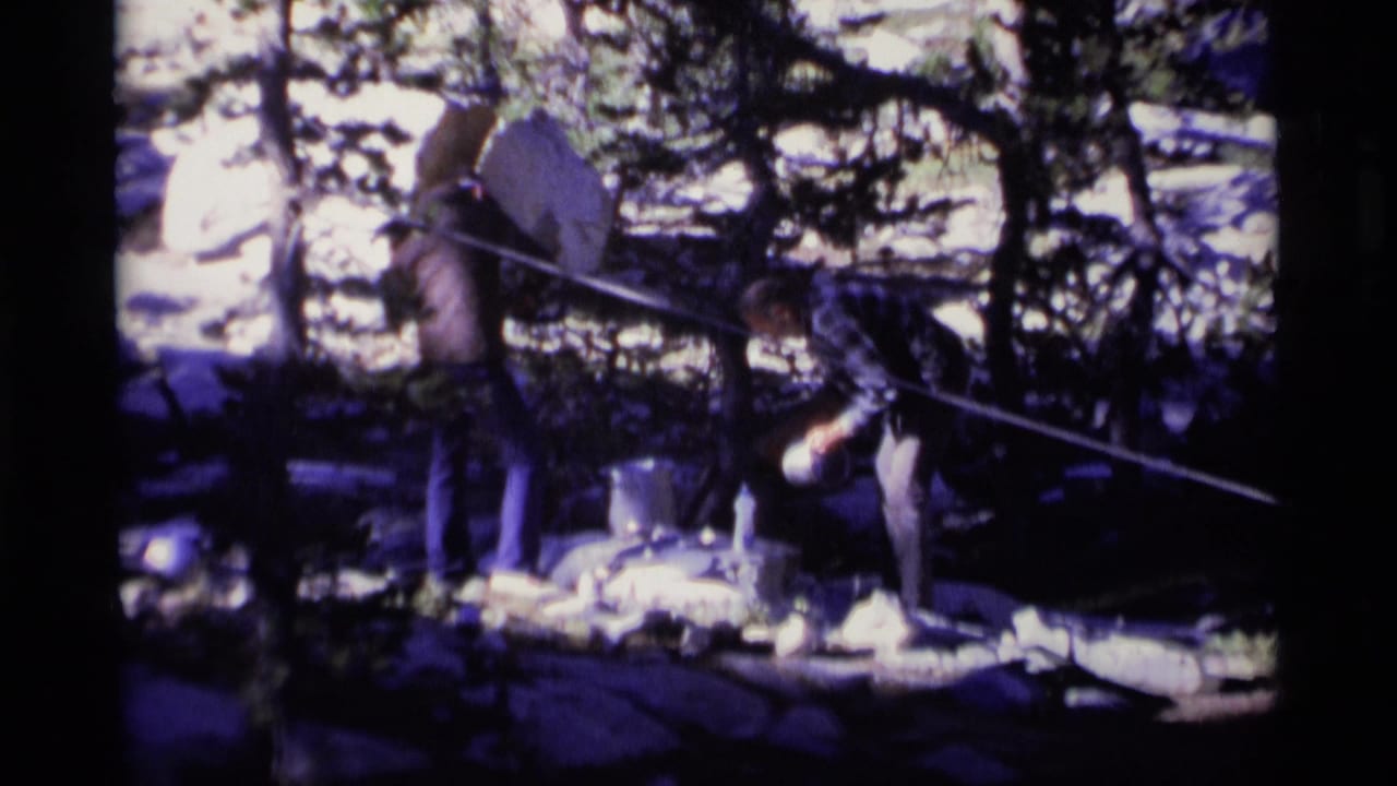 Two men at a wooded campsite with cookware and a rope line in front of them.