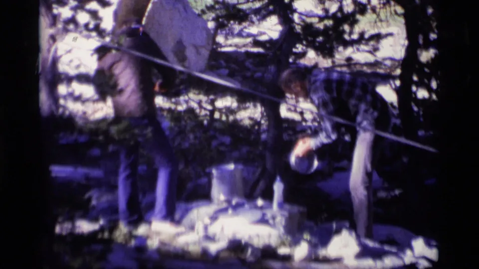 1975 Minaret Wilderness home-movie frame of a man bending toward cookware as another man stands in a wooded campsite.