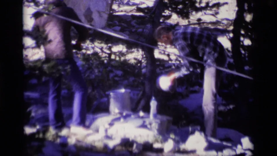 1975 Minaret Wilderness frame of a plaid-shirted man leaning over a pot with another adult nearby in a forest camp.