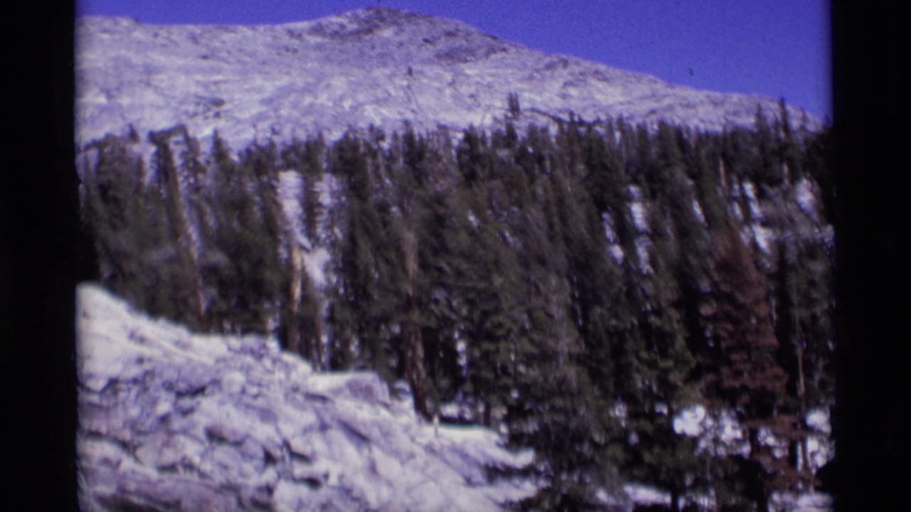 1975 8mm home-movie frame of Minaret Wilderness terrain with snow, pines, rocks, and a mountain ridge.