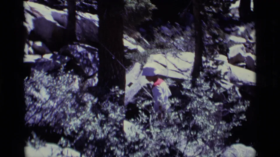 Hiker in a white hat and light clothing navigates dense brush and rocks in 1975 Minaret Wilderness 8mm footage.