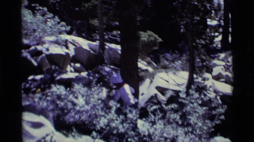 Hiker partially obscured by brush crosses rocky forest terrain in the Minaret Wilderness, captured on handheld 8mm film.