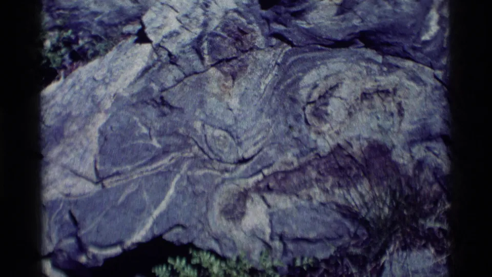 1975 8mm Minaret Wilderness frame showing a close boulder surface with branching cracks, layered stone, and a small patch of vegetation.