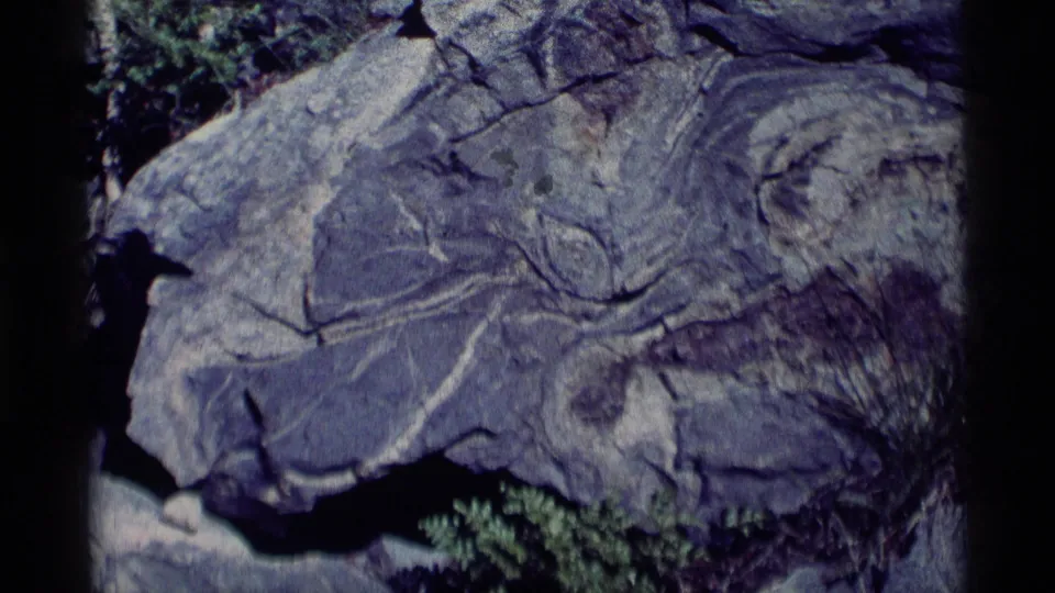 1975 8mm Minaret Wilderness close-up of an angled gray rock face with fissures, rough edges, and small green plants at the base.