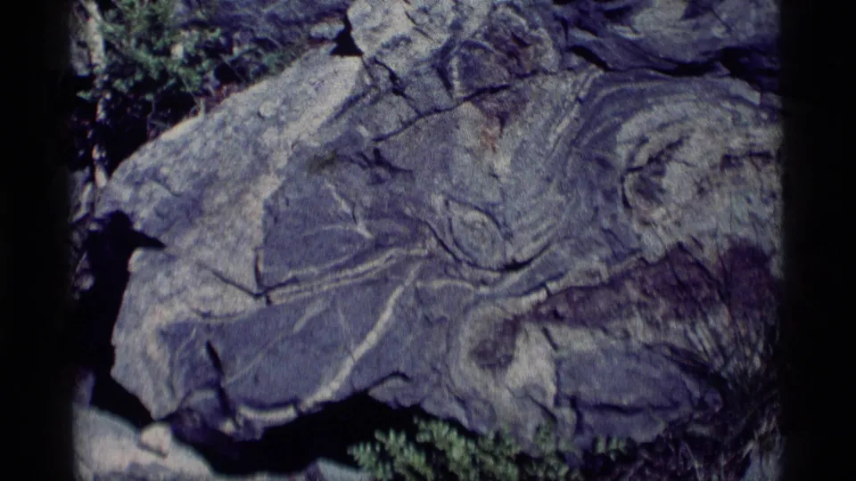 1975 8mm Minaret Wilderness view of a broad cracked boulder, layered stone markings, dark shadows, and green vegetation beneath.