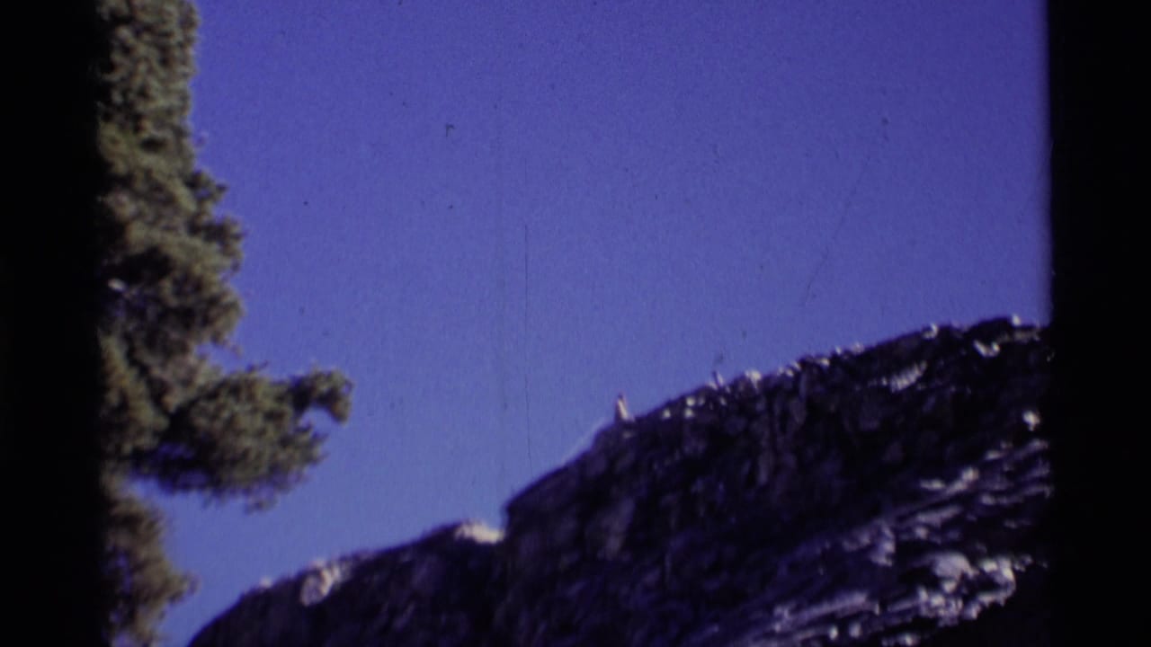1975 8mm frame from Minaret Wilderness showing a dark rocky ridge, blue sky and a conifer along the left edge.