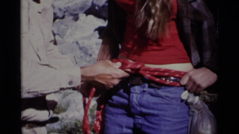 The red rope appears secured around the woman's waist while a pale-sleeved person makes a final adjustment in the 1975 wilderness footage.