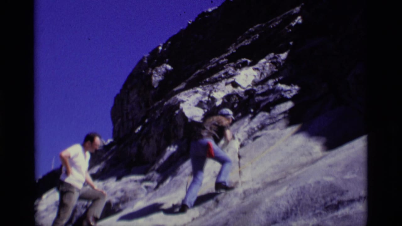 Two hikers on a steep rocky slope, one climbing with a rope while another waits below.