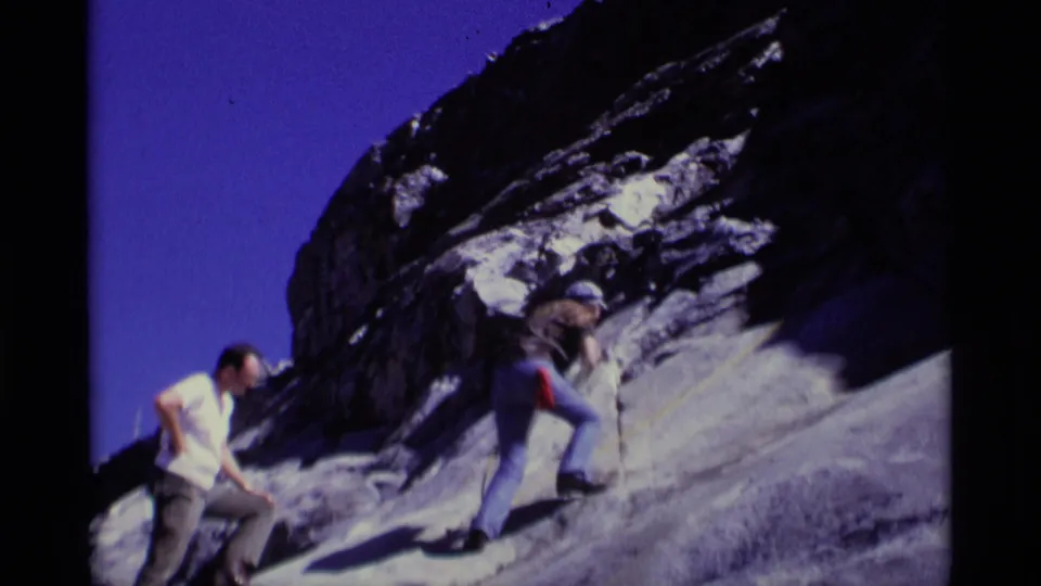 1975 Minaret Wilderness home-movie frame showing two adult hikers on a steep rock face, with one climber using a yellow rope.