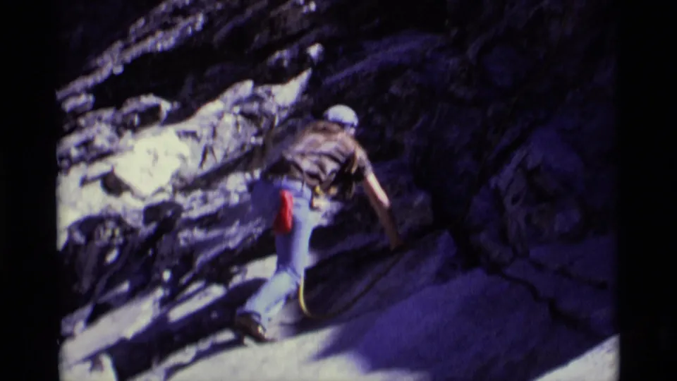 Minaret Wilderness home-movie frame shows the climber from behind traversing dark rock with rope and red gear visible.