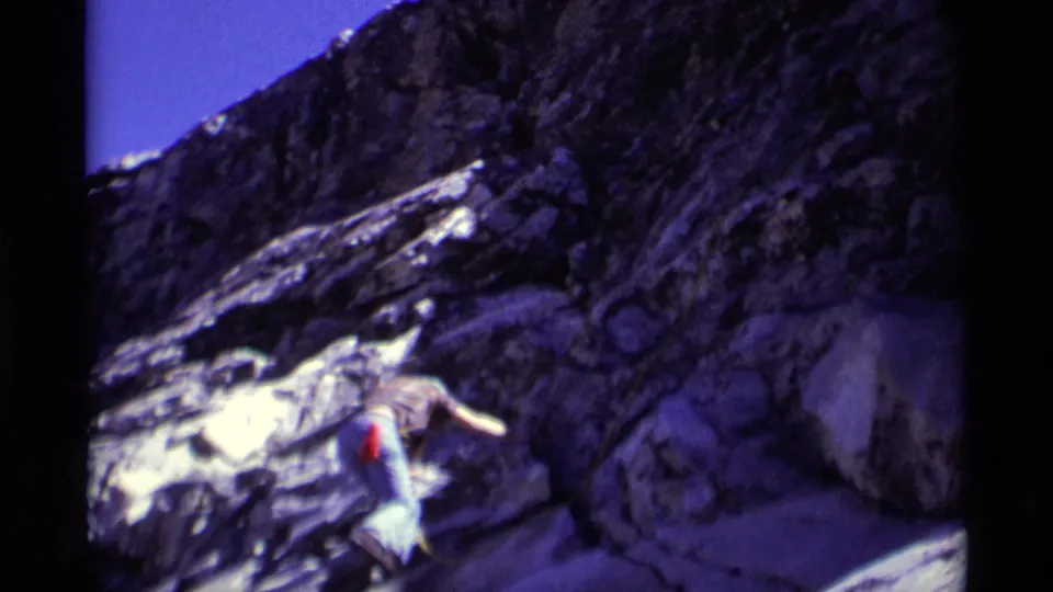 Wide 1975 8mm view of a climber on steep Minaret Wilderness rock, with dark cliffs and bright sky above.
