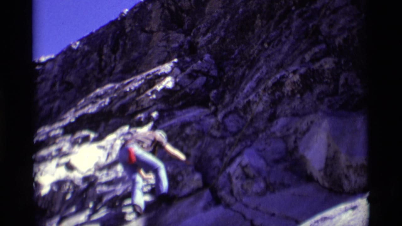 Climber in a white helmet moves across a steep dark rock face under blue sky.