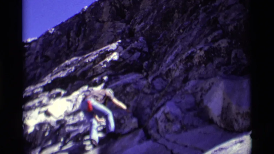 1975 Minaret Wilderness home-movie frame showing a helmeted climber traversing rugged rock with pale patches nearby.