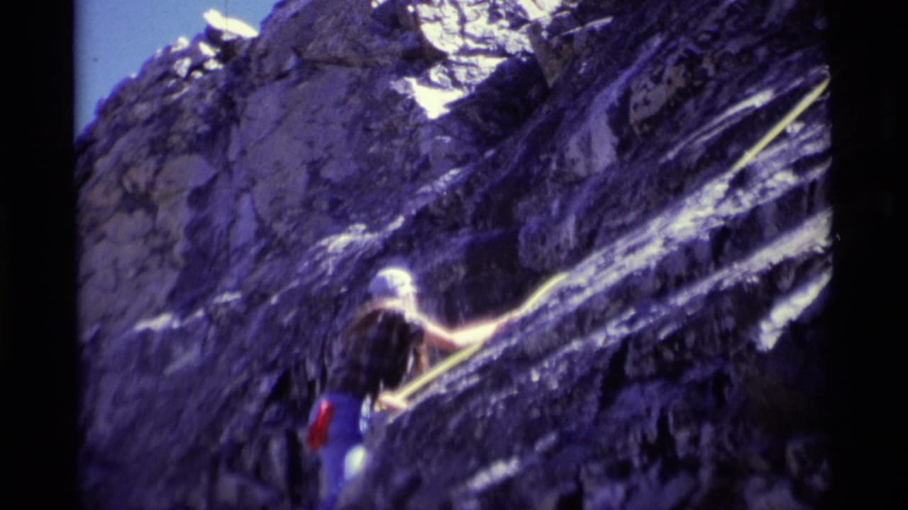 In 1975 Minaret Wilderness footage, a helmeted climber begins ascending a steep purple-toned rock face with a yellow rope.