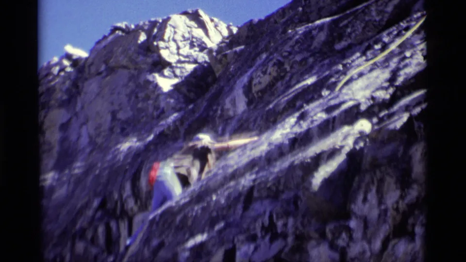 A helmeted climber leans into the steep Minaret Wilderness rock surface, continuing upward with rope support in grainy 8mm footage.