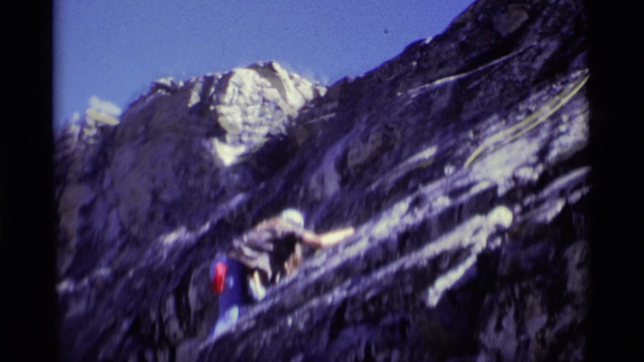 1975 8mm frame from Minaret Wilderness showing a climber with a red backpack on a steep rock face under a blue sky.