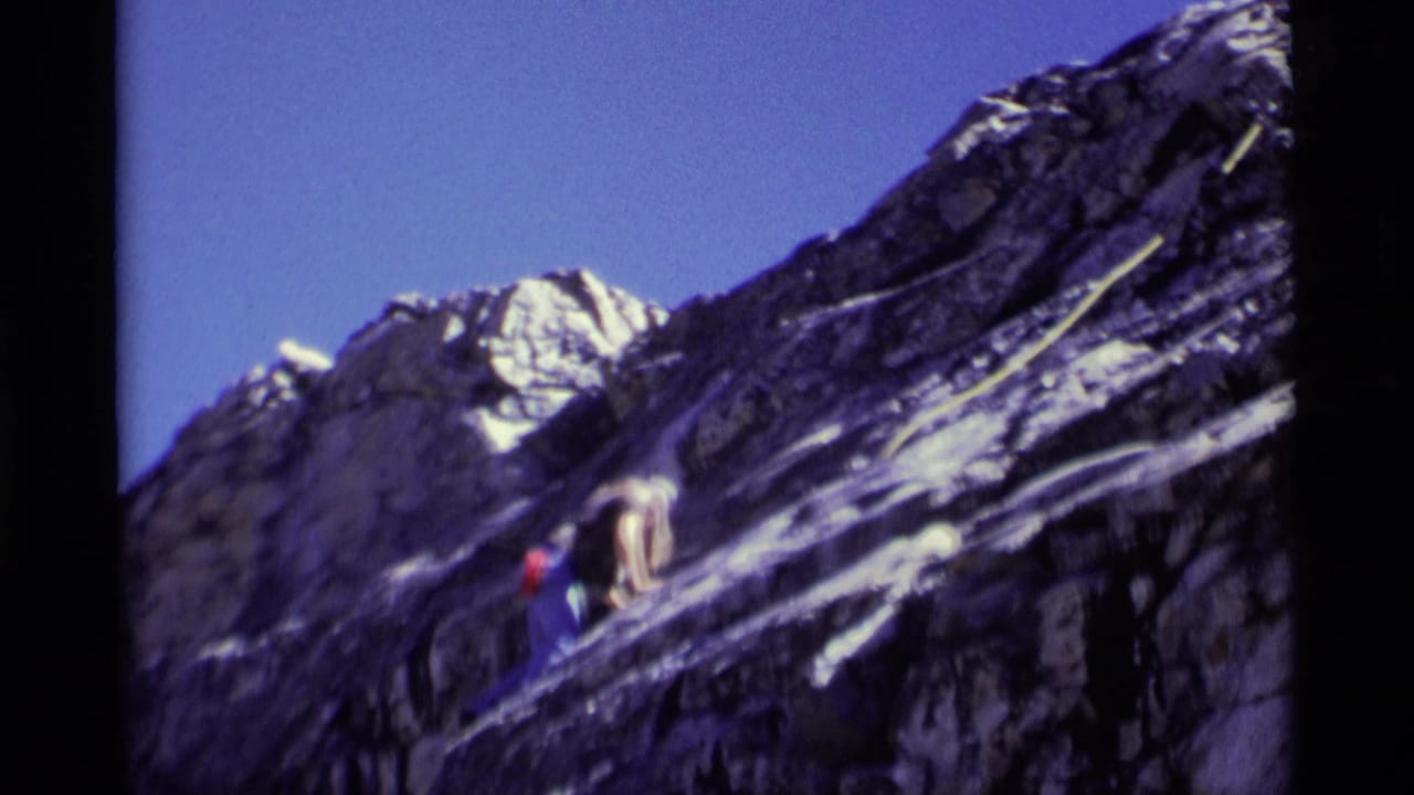 1975 Minaret Wilderness home-movie frame showing a climber ascending dark striated rock with a pale rope nearby.