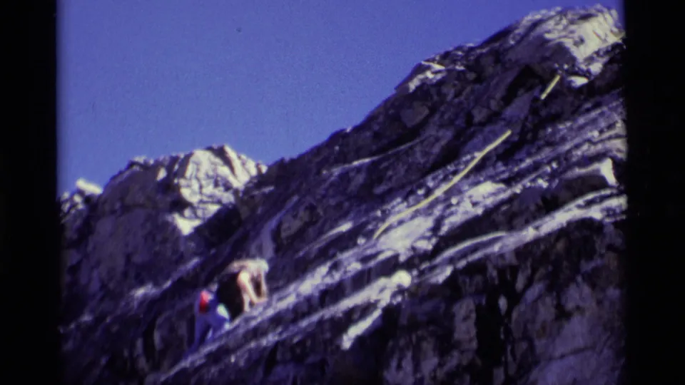 1975 8mm Minaret Wilderness frame with a climber crouched low on a steep rocky incline and a rope crossing the scene.