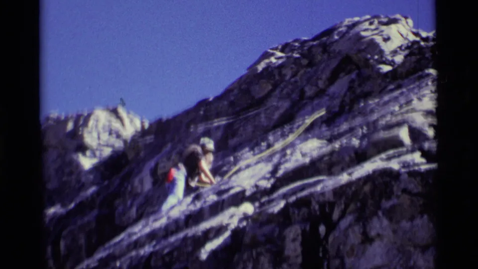 1975 Minaret Wilderness frame showing a climber beside a yellow rope on steep rock with snow-streaked peaks in the background.