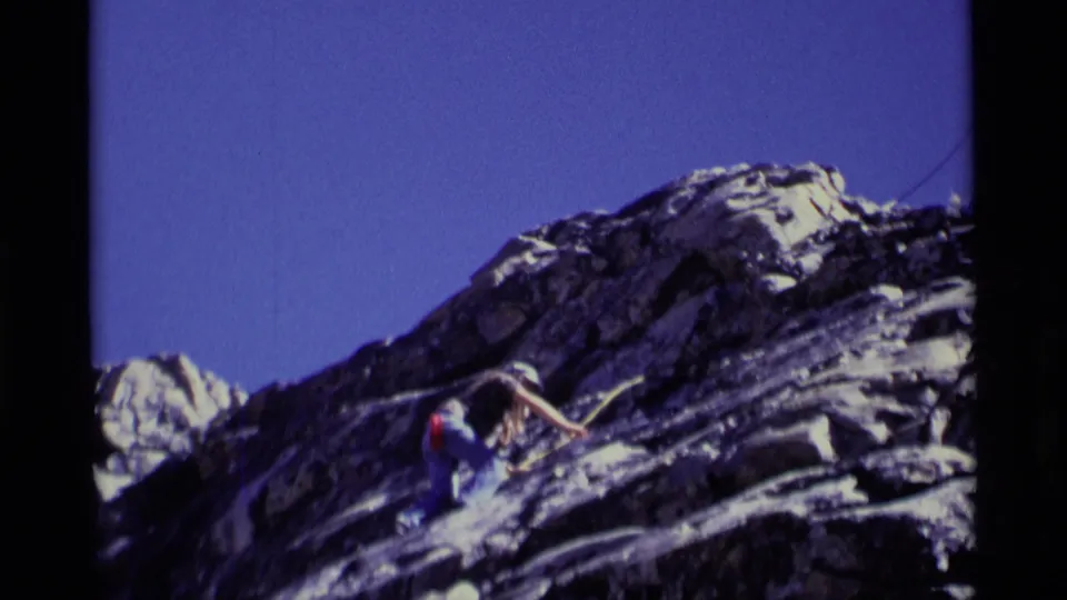A roped climber in light upper clothing and blue pants leans into the steep California wilderness rock face during a careful ascent.