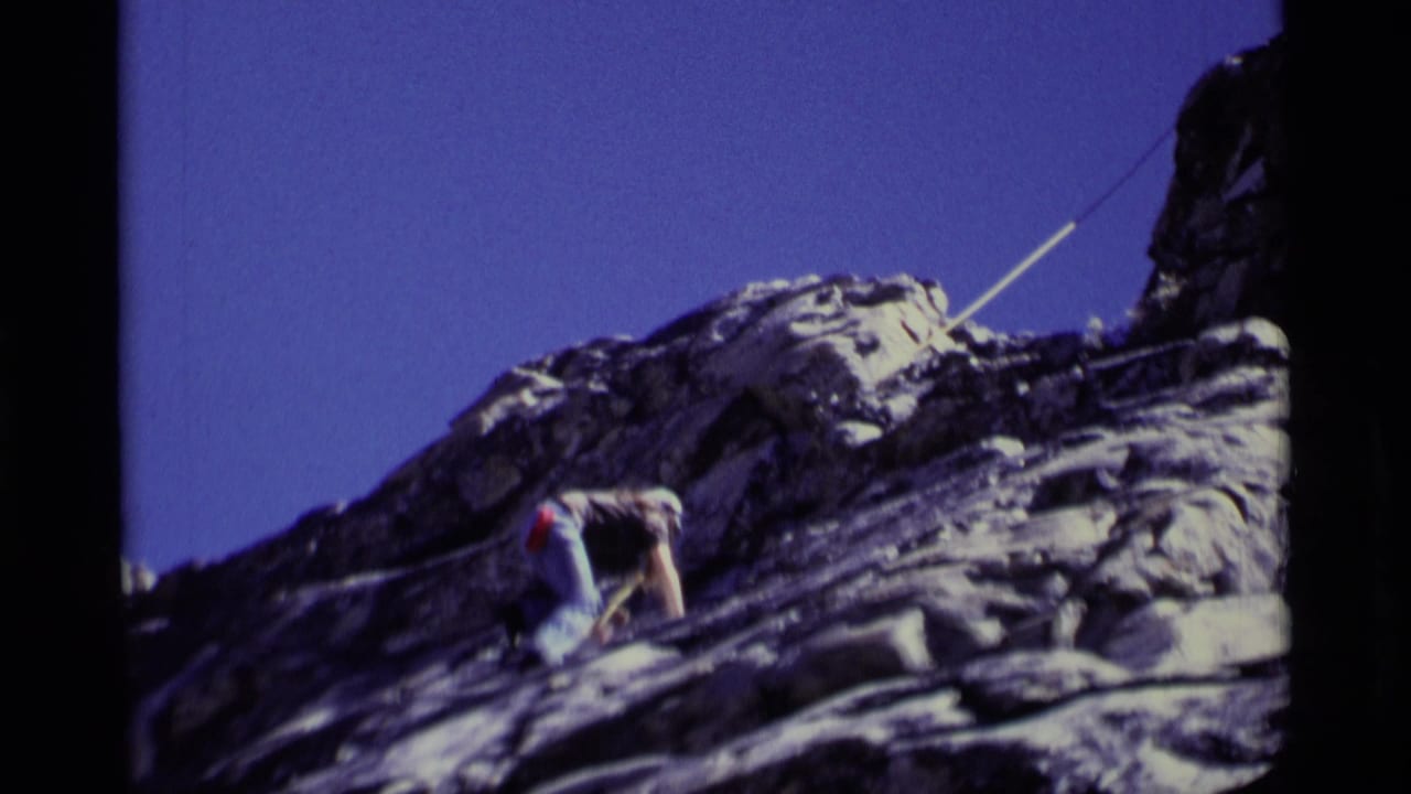 Climber attached to a rope moves across a steep gray rock face under blue sky.