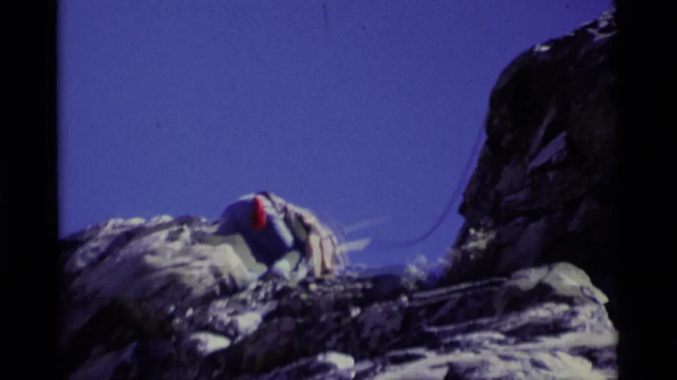 Roped climber in 1975 California wilderness footage bends over a rocky ledge, framed by blue sky and dark cliff shadow.