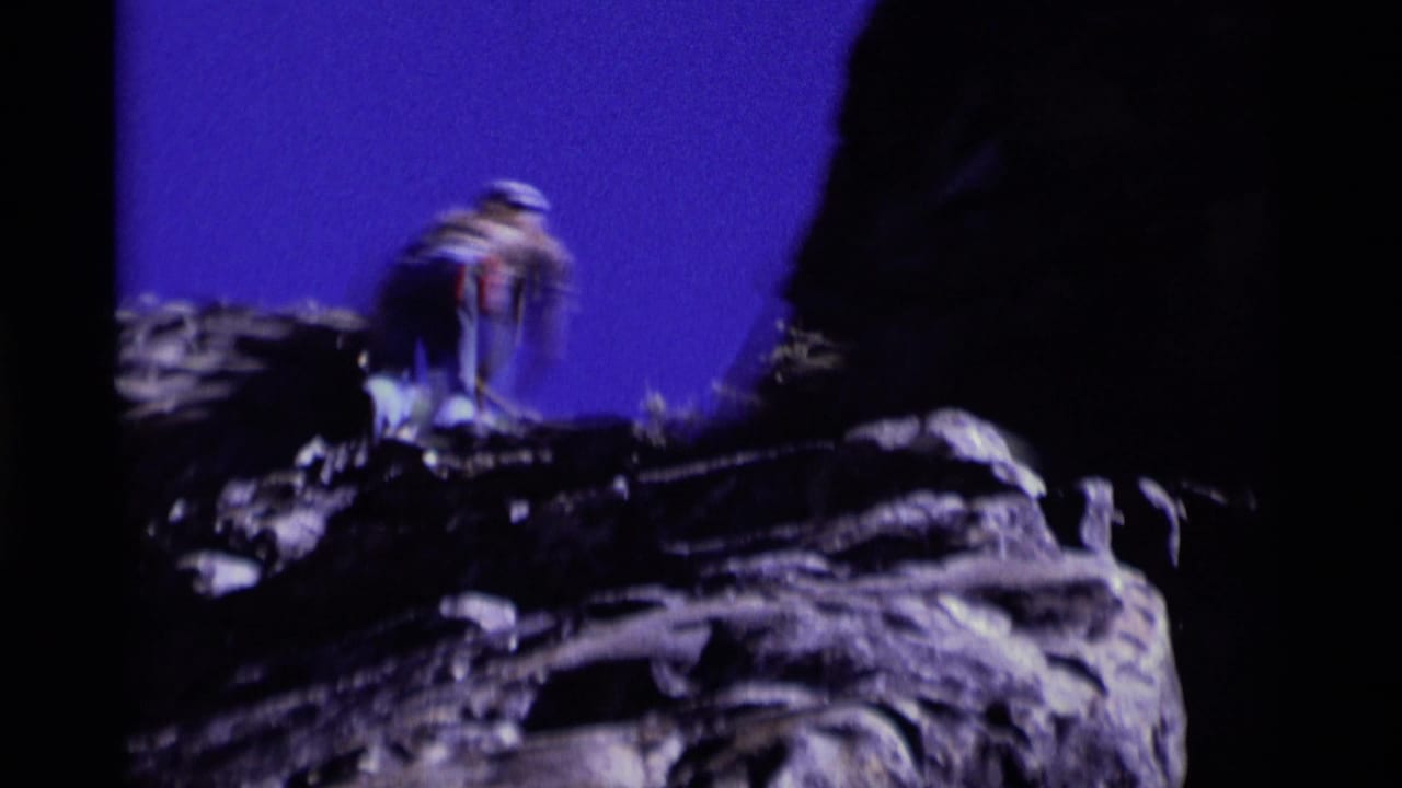 Roped climber in hat and jeans stands on a rugged Minaret Wilderness rock outcrop in a 1975 8mm home-movie frame.