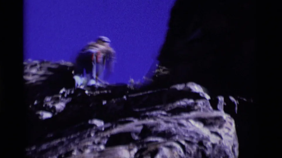 Roped climber in hat and jeans stands on a rugged Minaret Wilderness rock outcrop in a 1975 8mm home-movie frame.