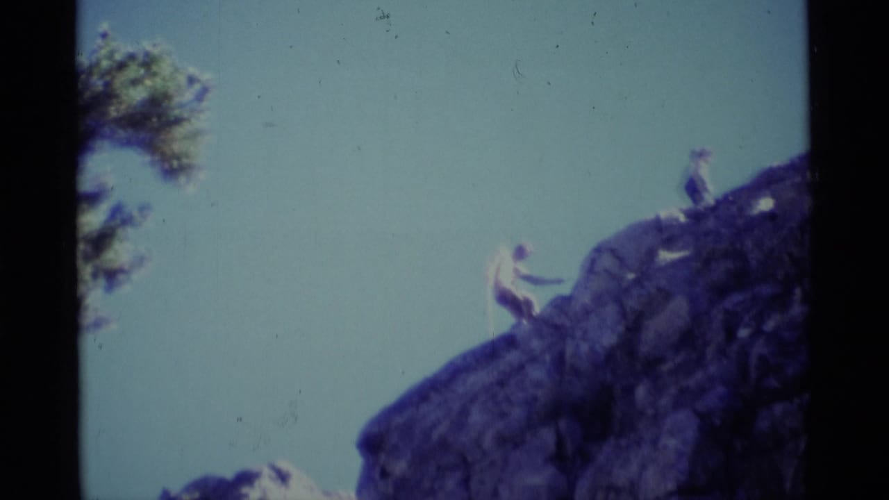 Two distant hikers on a steep rocky slope under a pale sky, with a tree branch at left.