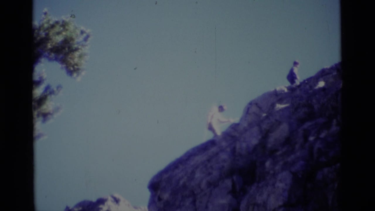 Distant 1975 California wilderness frame with one hiker on the sloped rock and another figure near the top of the formation.