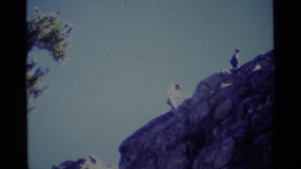 Distant 1975 California wilderness frame with one hiker on the sloped rock and another figure near the top of the formation.
