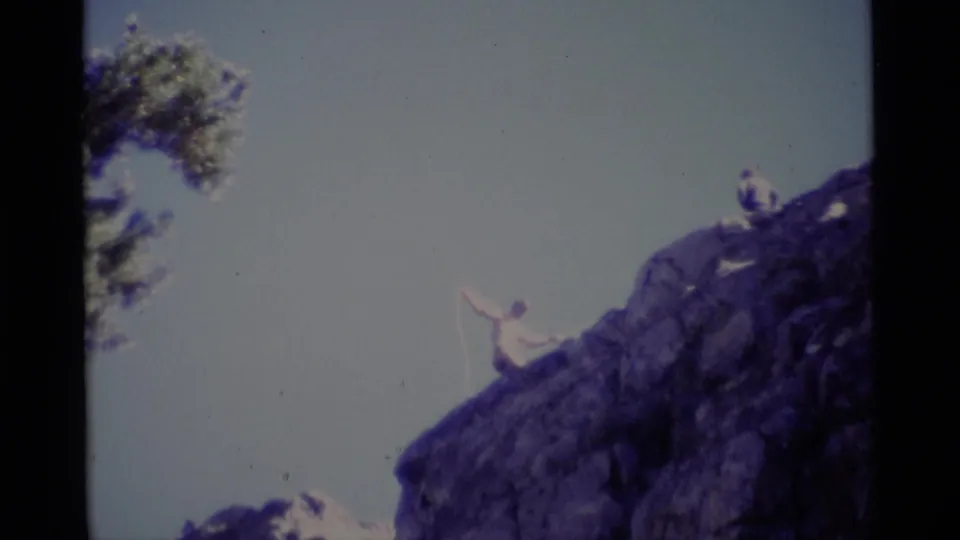 1975 8mm frame showing distant hikers on exposed Minaret Wilderness rock, with one figure lifting an arm beside a rope-like line.