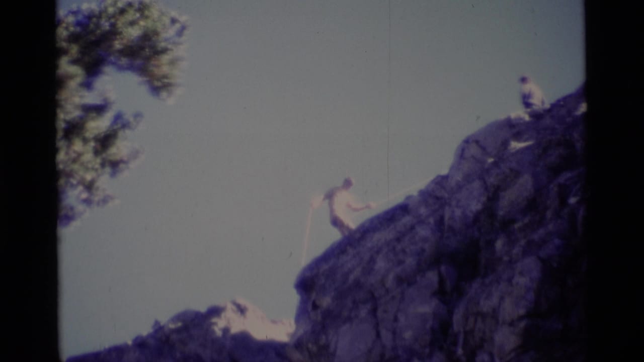 1975 California wilderness frame with a roped climber on dark rock and a second person at the summit edge.