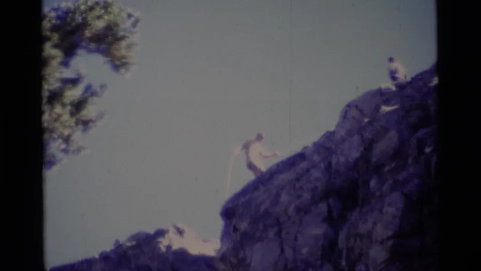 1975 California wilderness frame with a roped climber on dark rock and a second person at the summit edge.