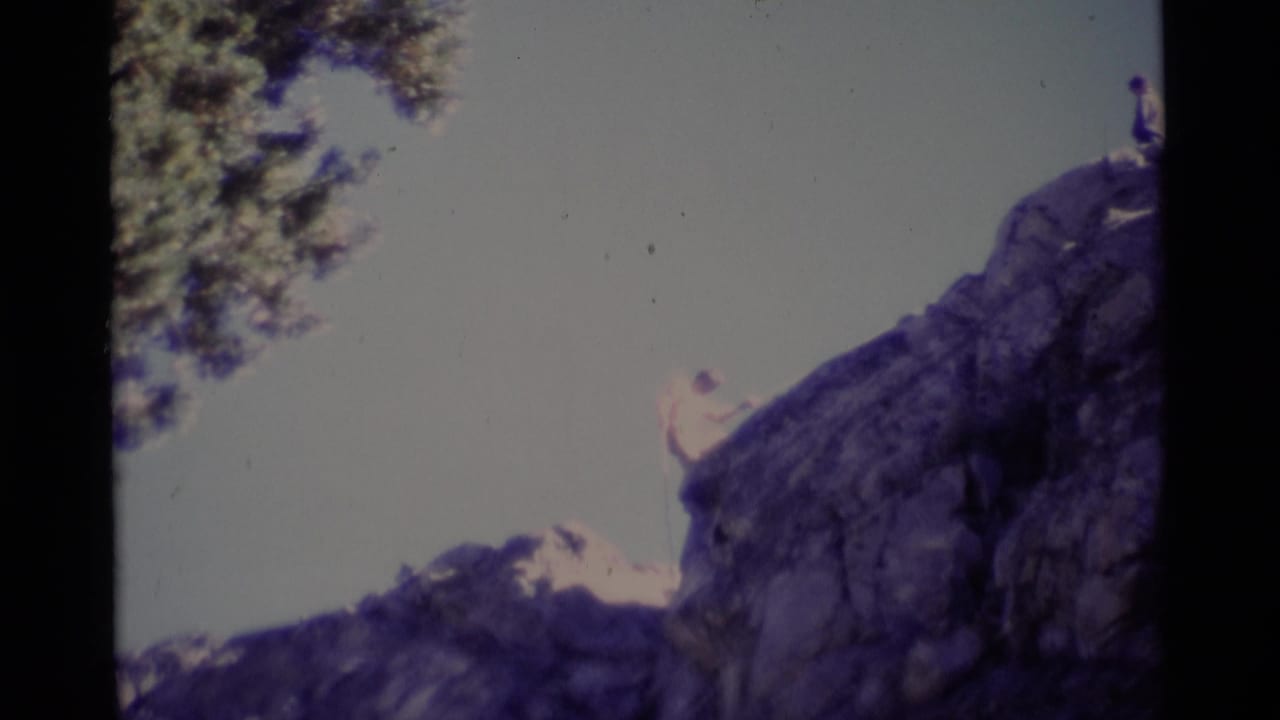 1975 Minaret Wilderness 8mm frame showing a man near the upper edge of a steep rock face with tree branches at left.