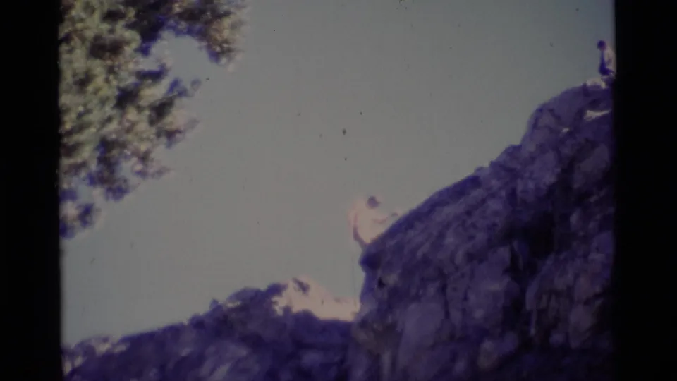 1975 Minaret Wilderness 8mm frame showing a man near the upper edge of a steep rock face with tree branches at left.