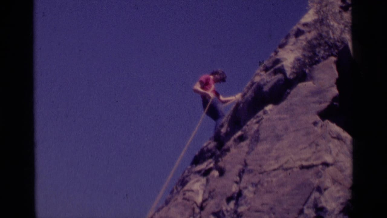 Climber in a red shirt ascends a steep rock face with a rope against blue sky.