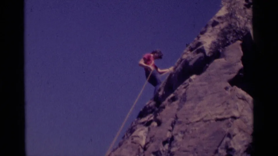 1975 8mm frame from Minaret Wilderness showing a climber in a red shirt moving up a steep rock face with a single rope.