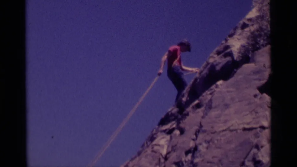 A red-shirted climber descends or shifts position on the steep Minaret Wilderness rock face, with rope visible against the sky.