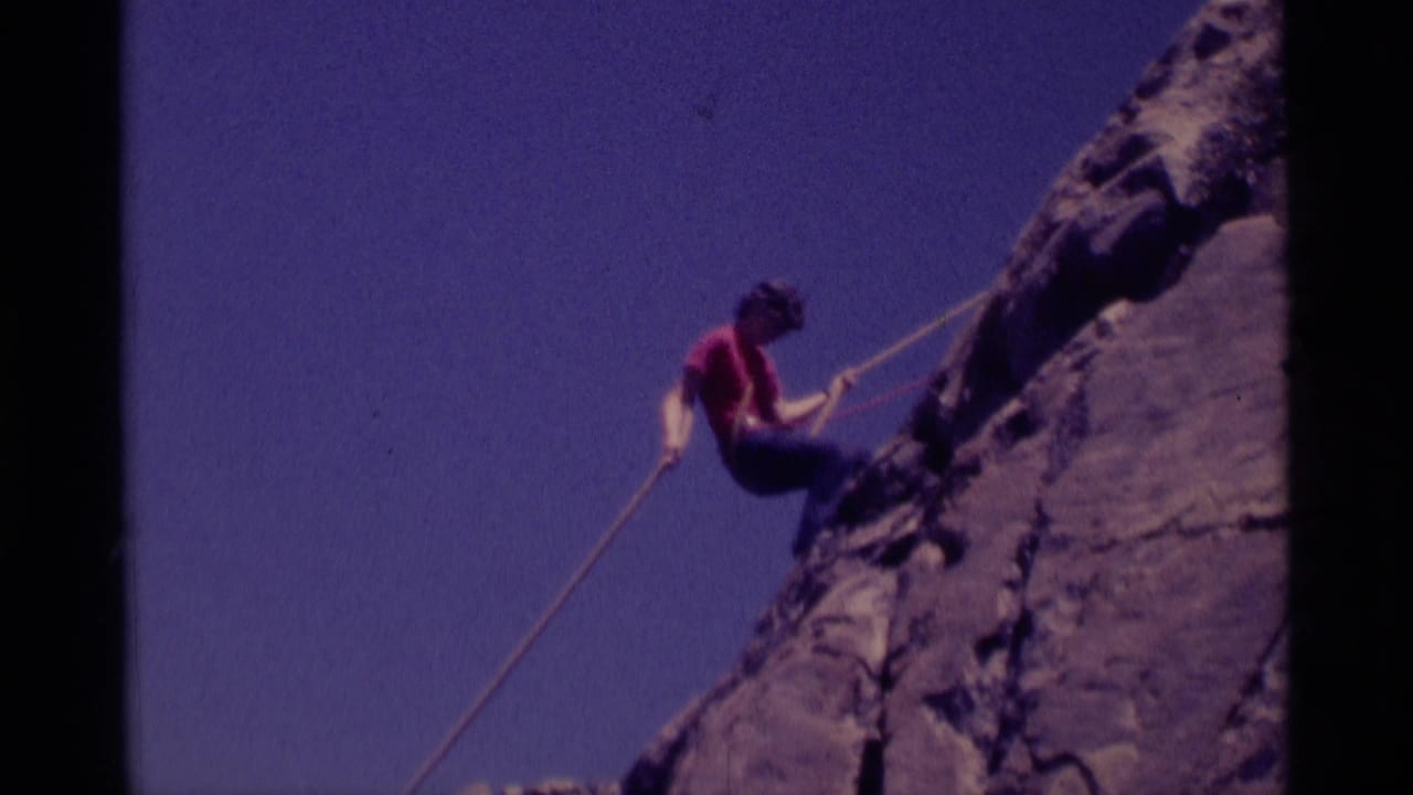 Climber in a red shirt rappels beside a steep rock face under a blue sky.
