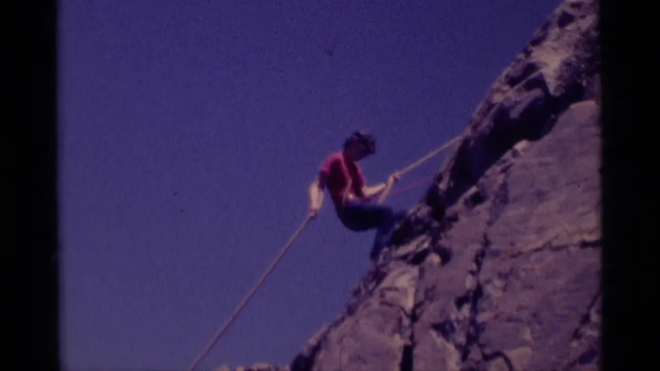 1975 8mm frame from Minaret Wilderness showing a roped climber in a red shirt descending along a rocky cliff under clear sky.