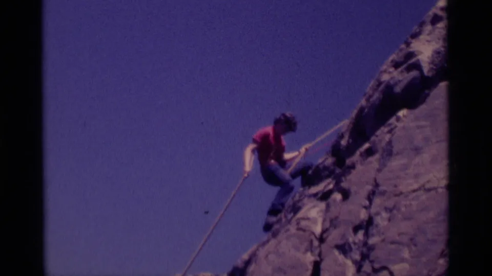 A climber in red continues rappelling down rock in 1975 Minaret Wilderness home-movie footage, framed against blue sky.