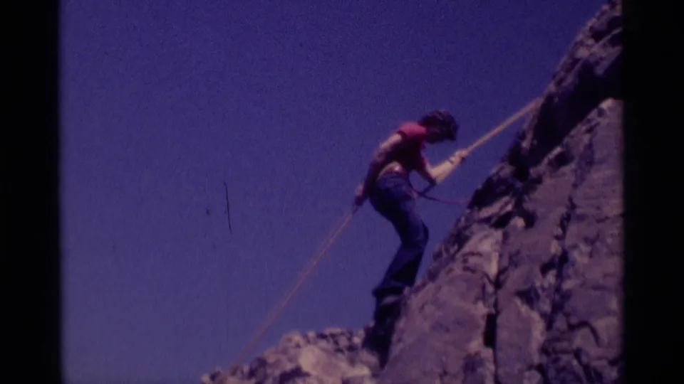 1975 8mm home-movie frame showing the rappeller partway down the cliff, with rope, rock texture, and sky visible.